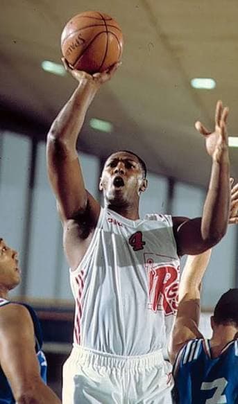Mike Mitchell con la maglia bianca numero 4 della Pallacanestro Reggiana, anni '90: tiro in sospensione in partita, avversari in maglia blu. Foto d'archivio trovata online, autore sconosciuto. Bassa risoluzione (343×583).